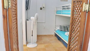 Una habitación con literas, un ventilador de pie, un televisor y una puerta con celosía decorativa.