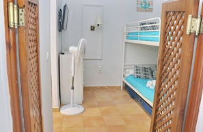 Una habitación con literas, un ventilador de pie, un televisor y una puerta con celosía decorativa.