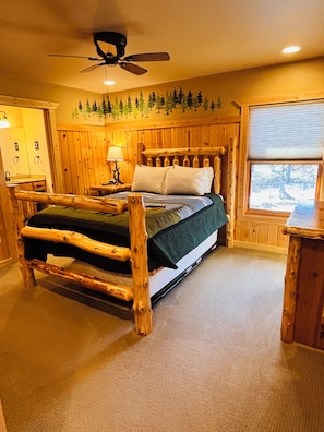 Ein Bett mit Holzrahmen, ein Deckenventilator, ein Nachttisch mit Lampe und ein Fenster mit Blick auf Bäume.