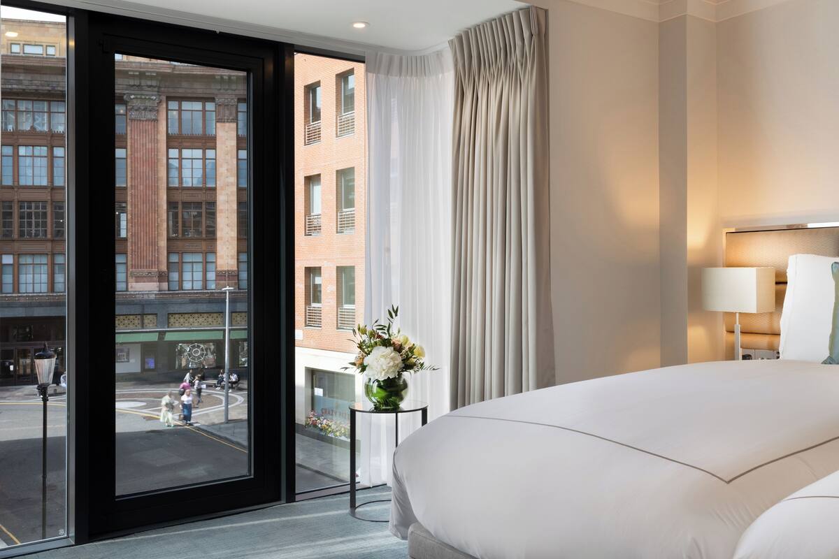 A hotel room with a large bed, a view of a city street, a vase with flowers, and a lamp on a bedside table.