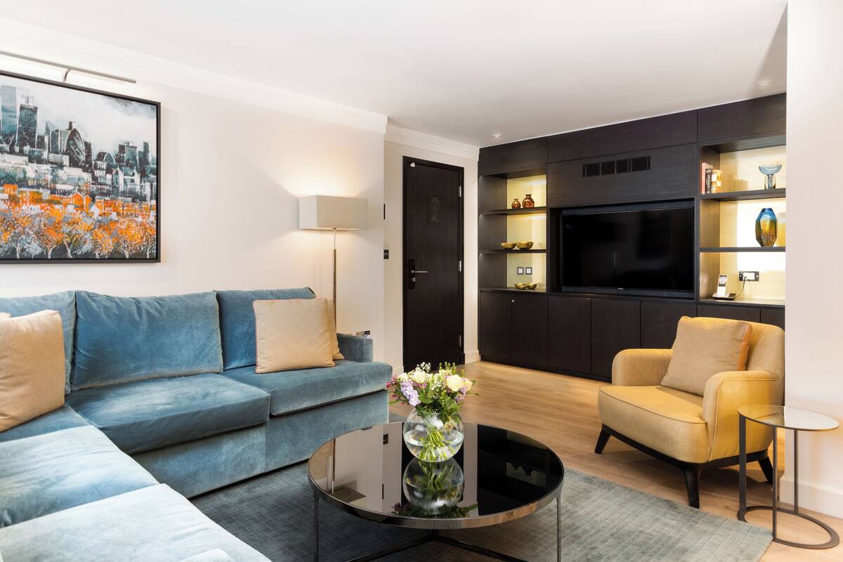 A modern living room with a sectional sofa, a round coffee table with a vase of flowers, a yellow armchair, a TV mounted on a dark cabinet, and a cityscape painting on the wall.