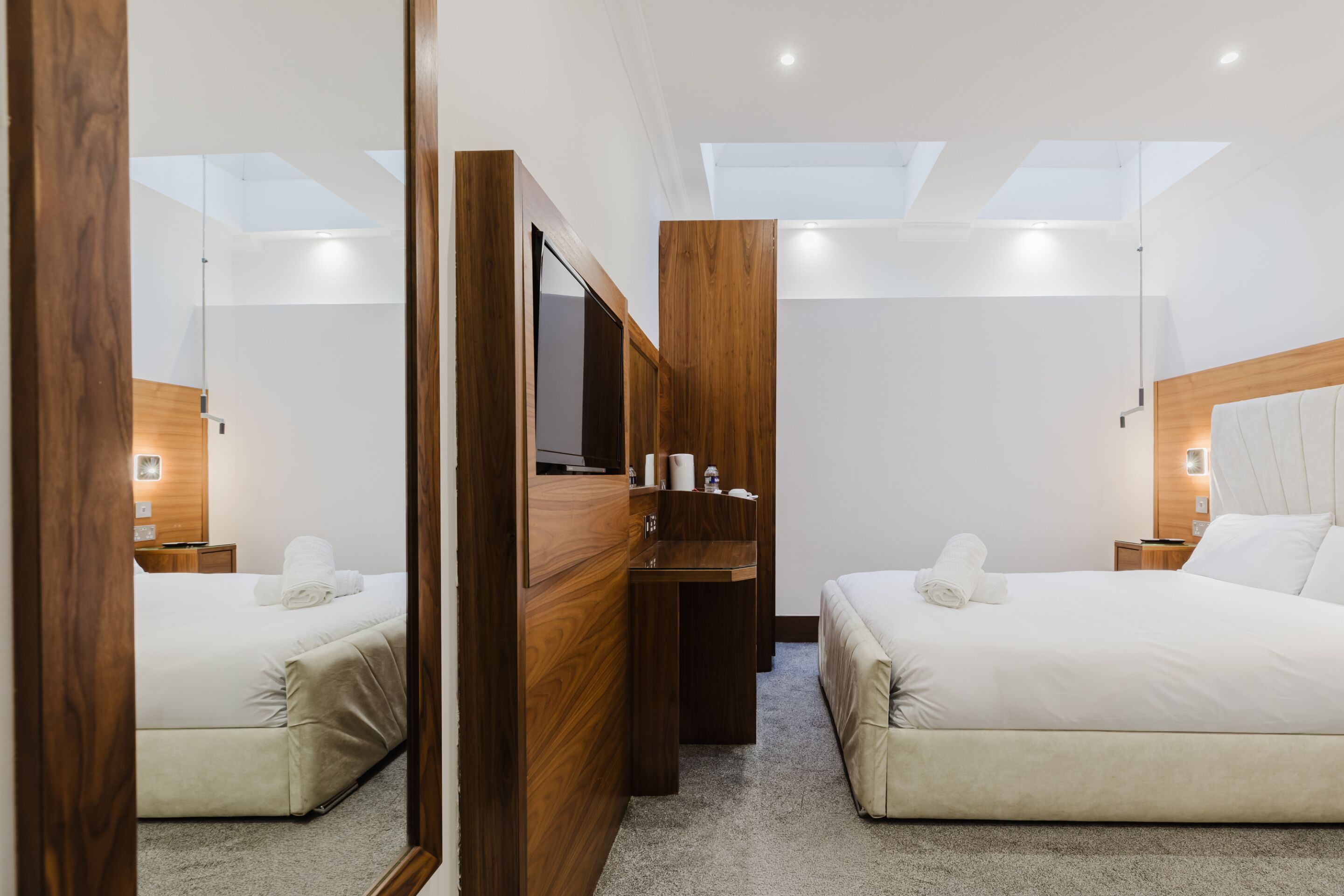 A hotel room with a large mirror, two beds, a wooden dresser, a small desk, and a skylight.