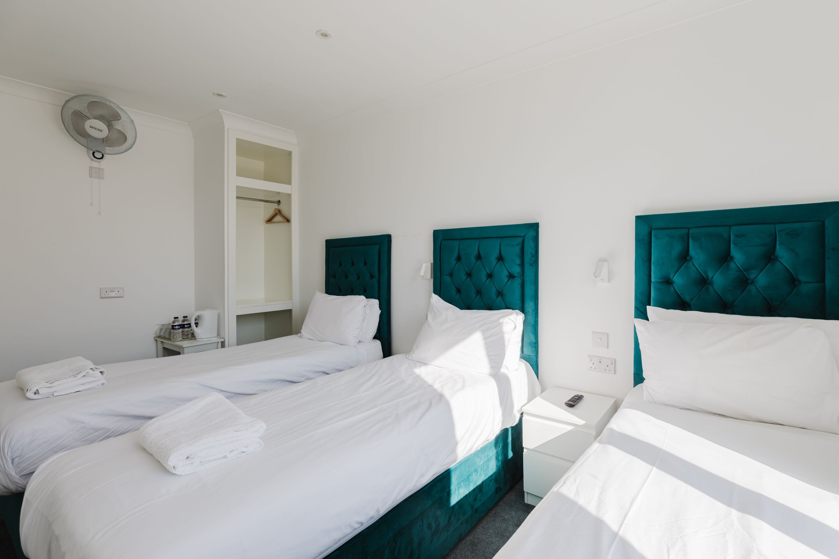 A hotel room with two beds, white linens, teal headboards, a bedside table with a phone, and a wall-mounted fan.