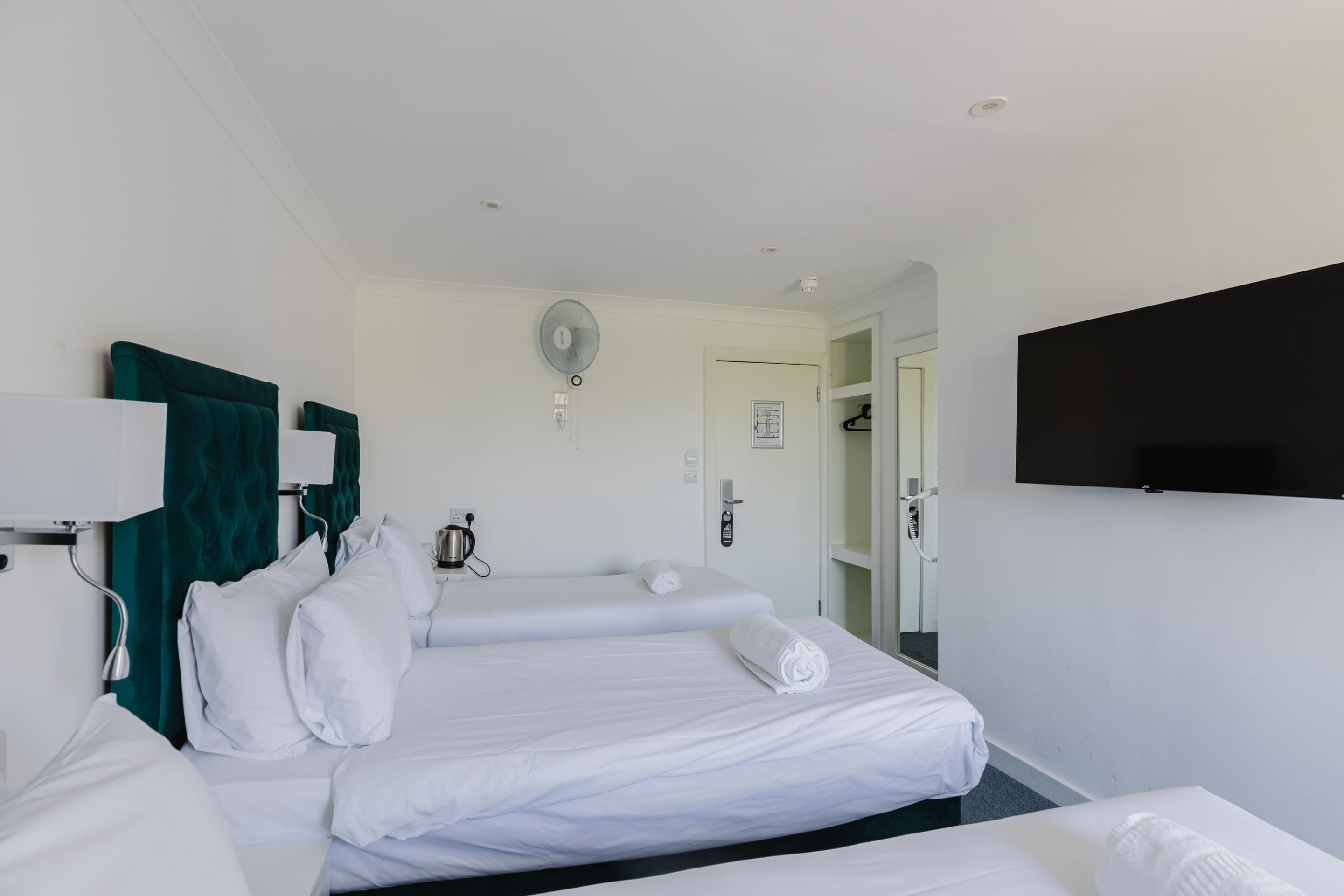 A hotel room with two beds, a flat-screen TV, a fan, and a door leading to a hallway.