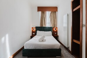 A neatly made bed with a green headboard, two bedside tables with lamps, a window with curtains, and a radiator. - The Gate Hotel (London)