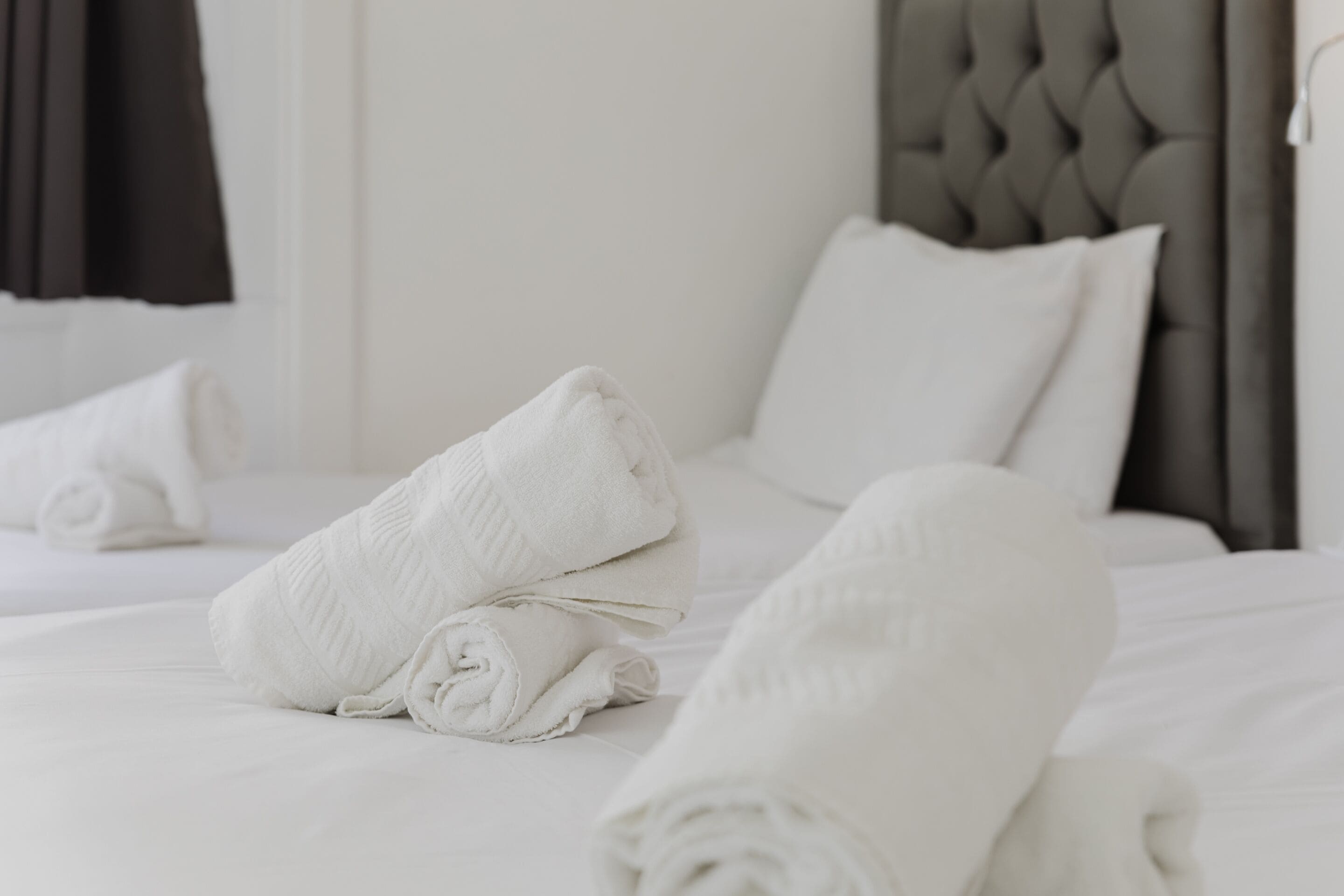 A neatly made bed with white linens and rolled towels, a tufted headboard, and a dark curtain.