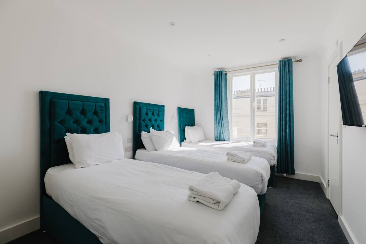 Une chambre d’hôtel avec trois lits, des têtes de lit turquoise, du linge de lit blanc et une fenêtre ornée de rideaux turquoise.