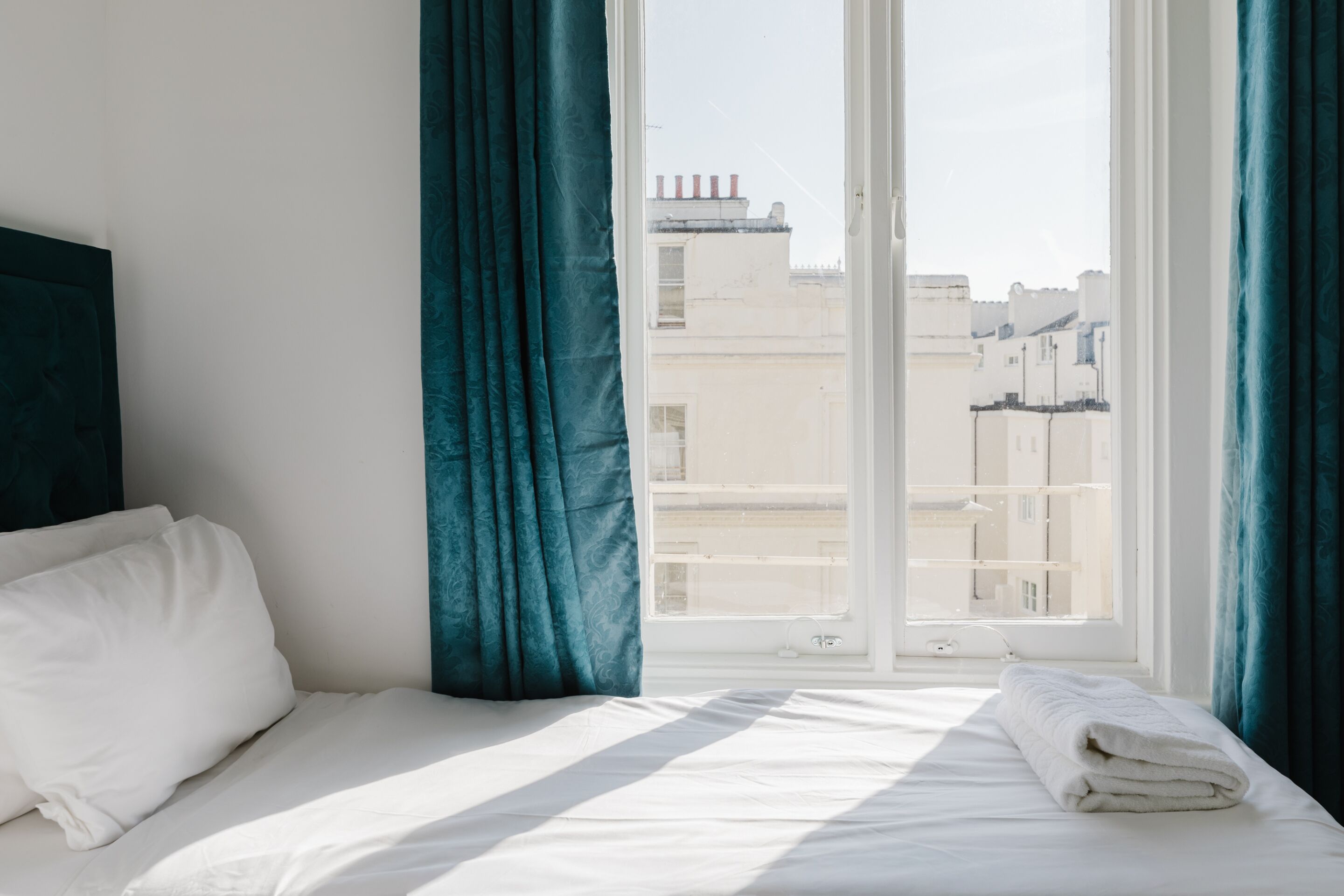 A hotel room with a bed, a window with curtains, and a view of buildings outside.