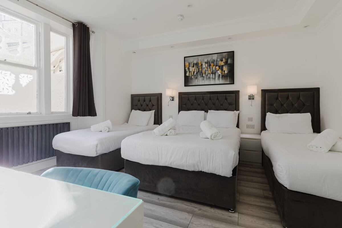Une chambre d’hôtel avec deux lits, un bureau, une chaise, une fenêtre avec des rideaux et un tableau au mur.