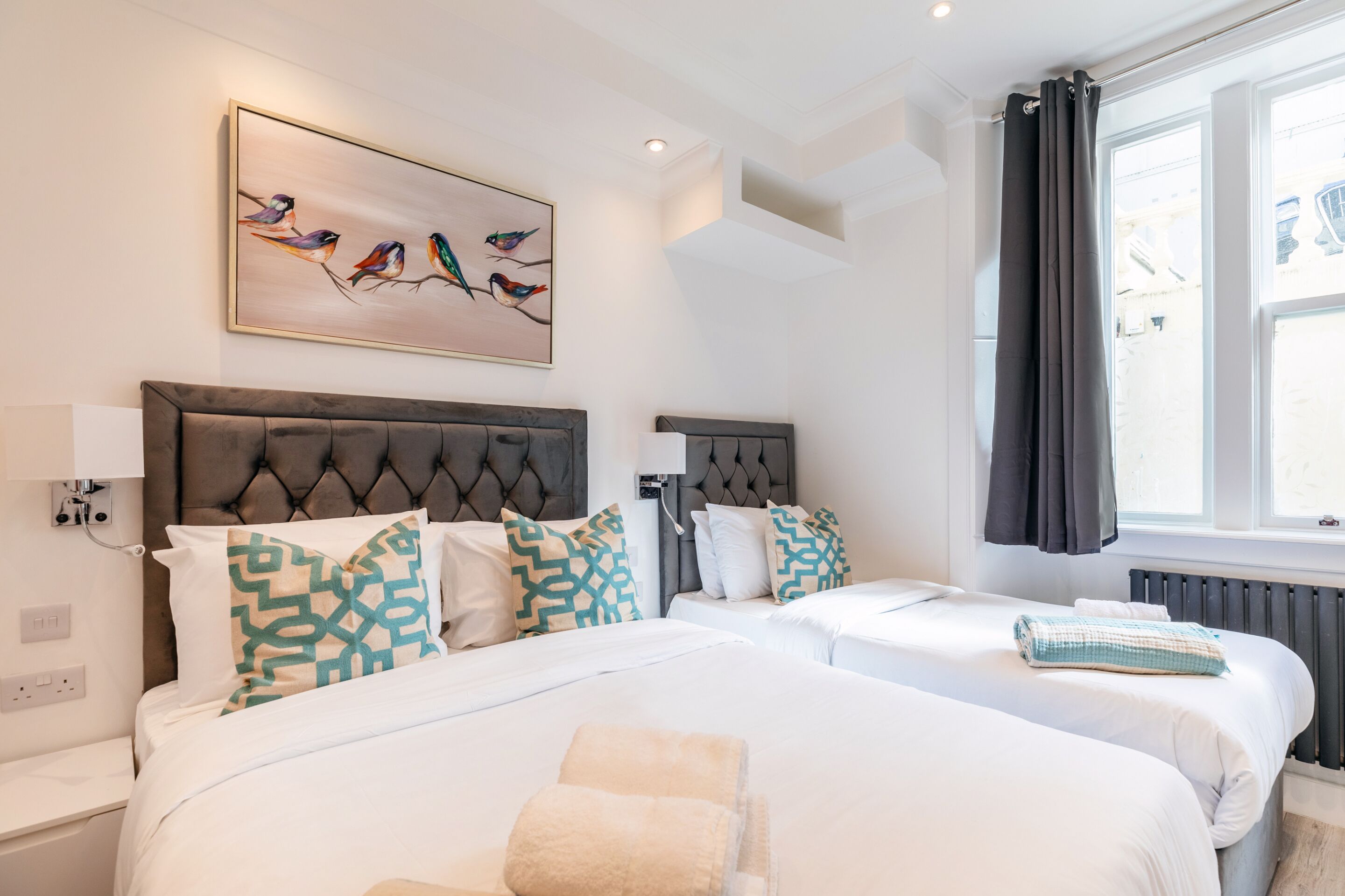 A hotel room with two beds, a large window with curtains, a wall-mounted painting, and a radiator.