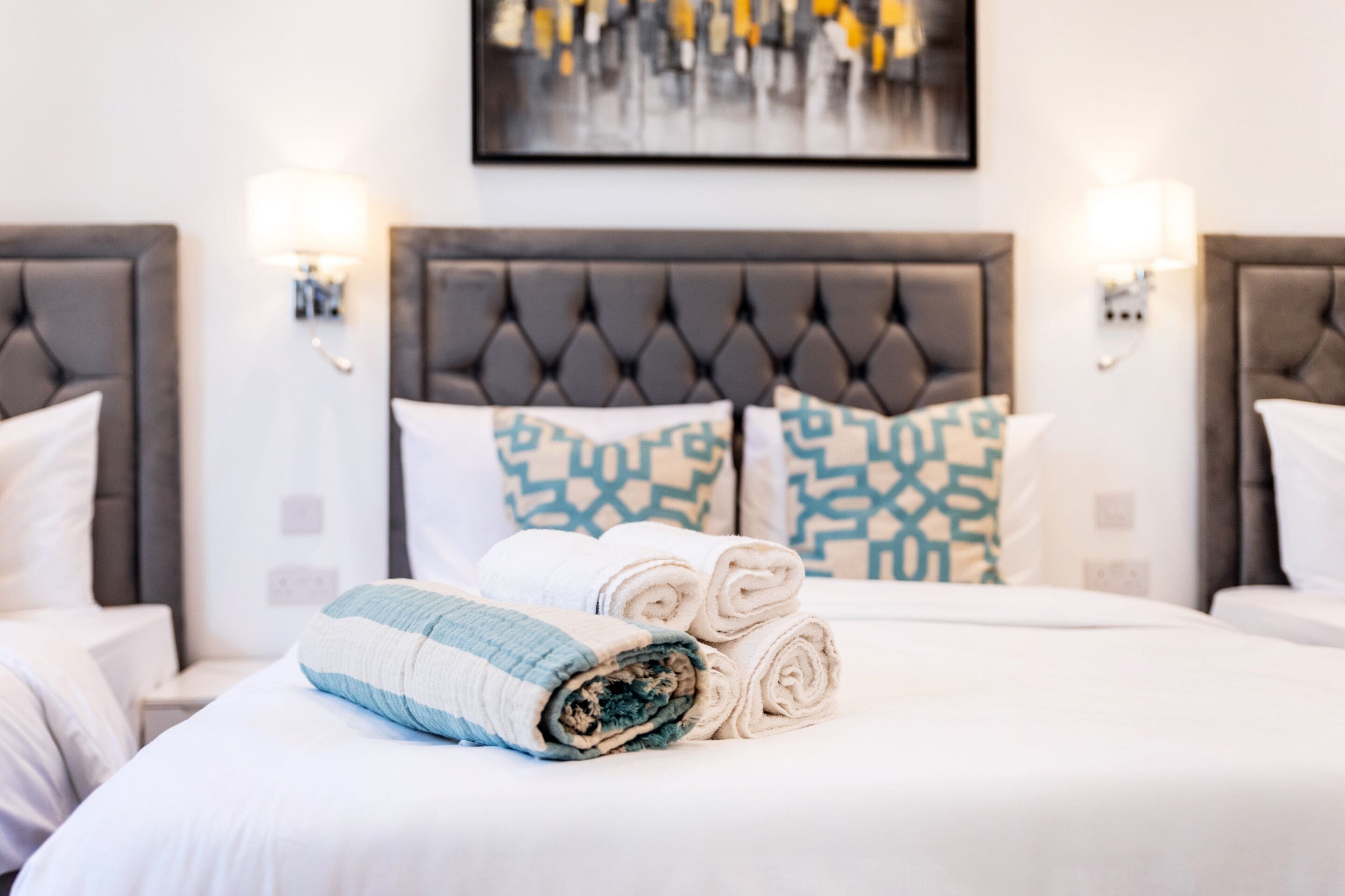 Habitación de hotel con dos camas, cada una con cabecero capitonado, ropa de cama blanca y almohadas estampadas. Hay dos lámparas de pared a cada lado de las camas.
