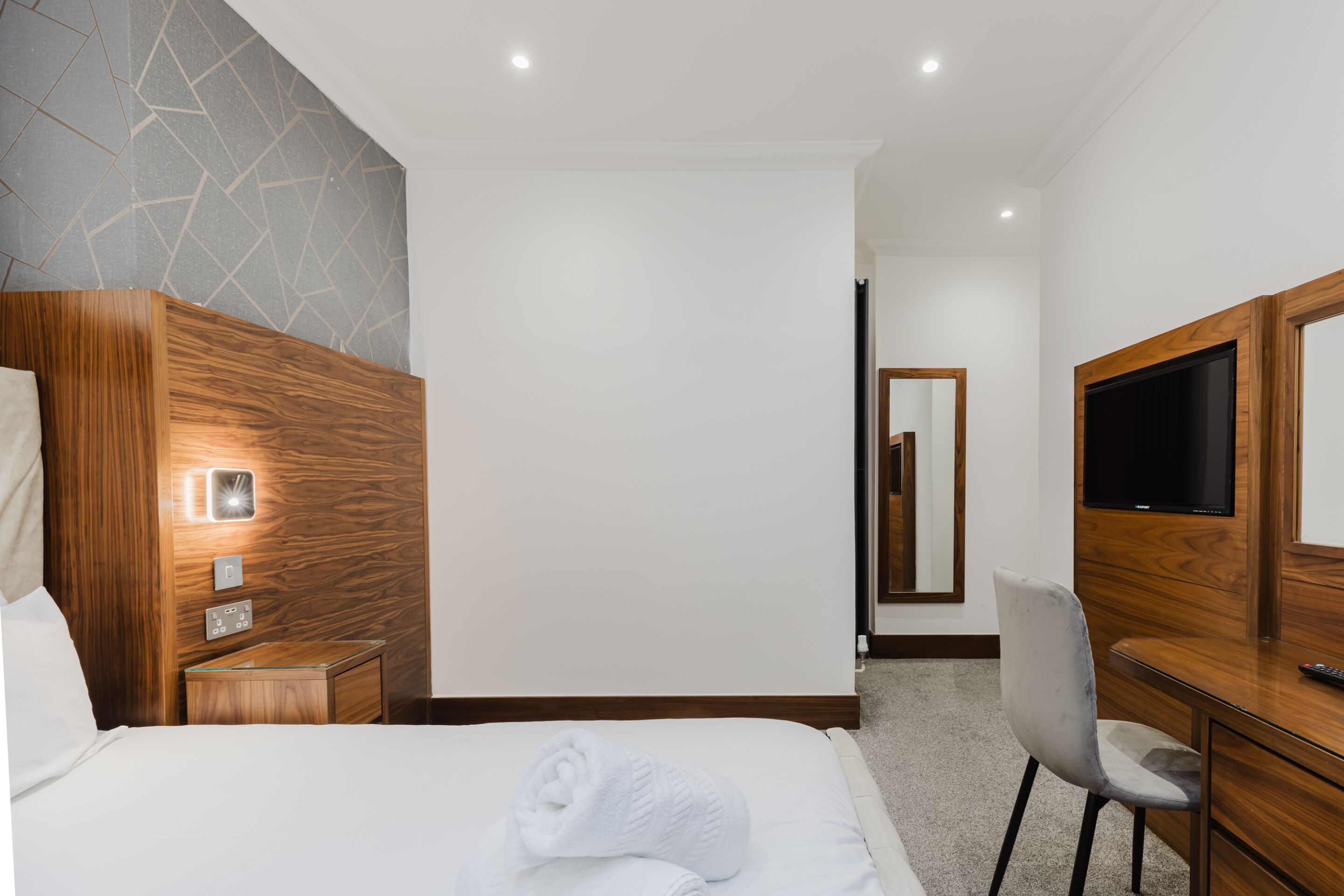 A hotel room with a bed, a wooden nightstand, a TV, a desk, and a chair.
