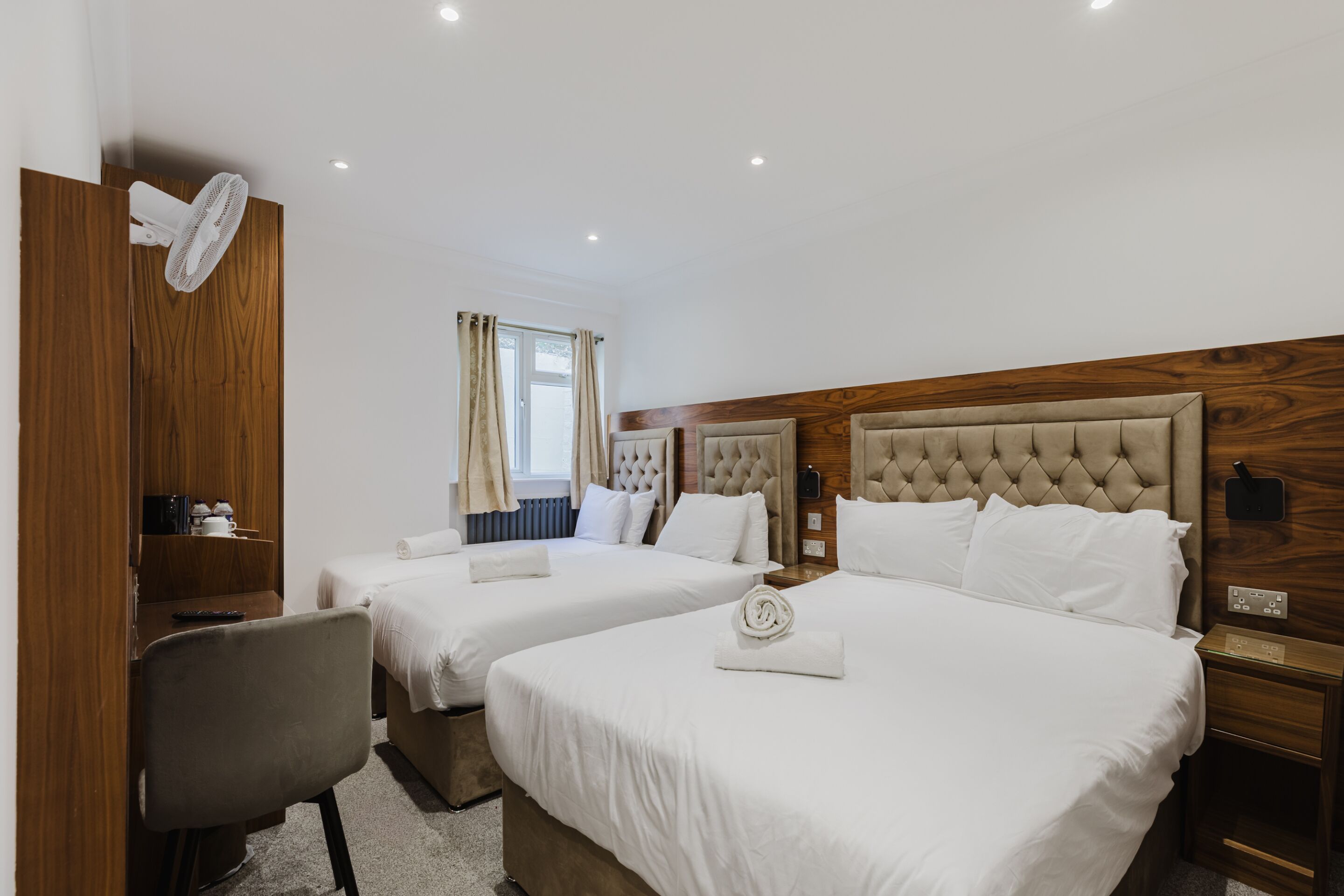 A hotel room with two beds, a desk, a chair, a wardrobe, and a ceiling fan.