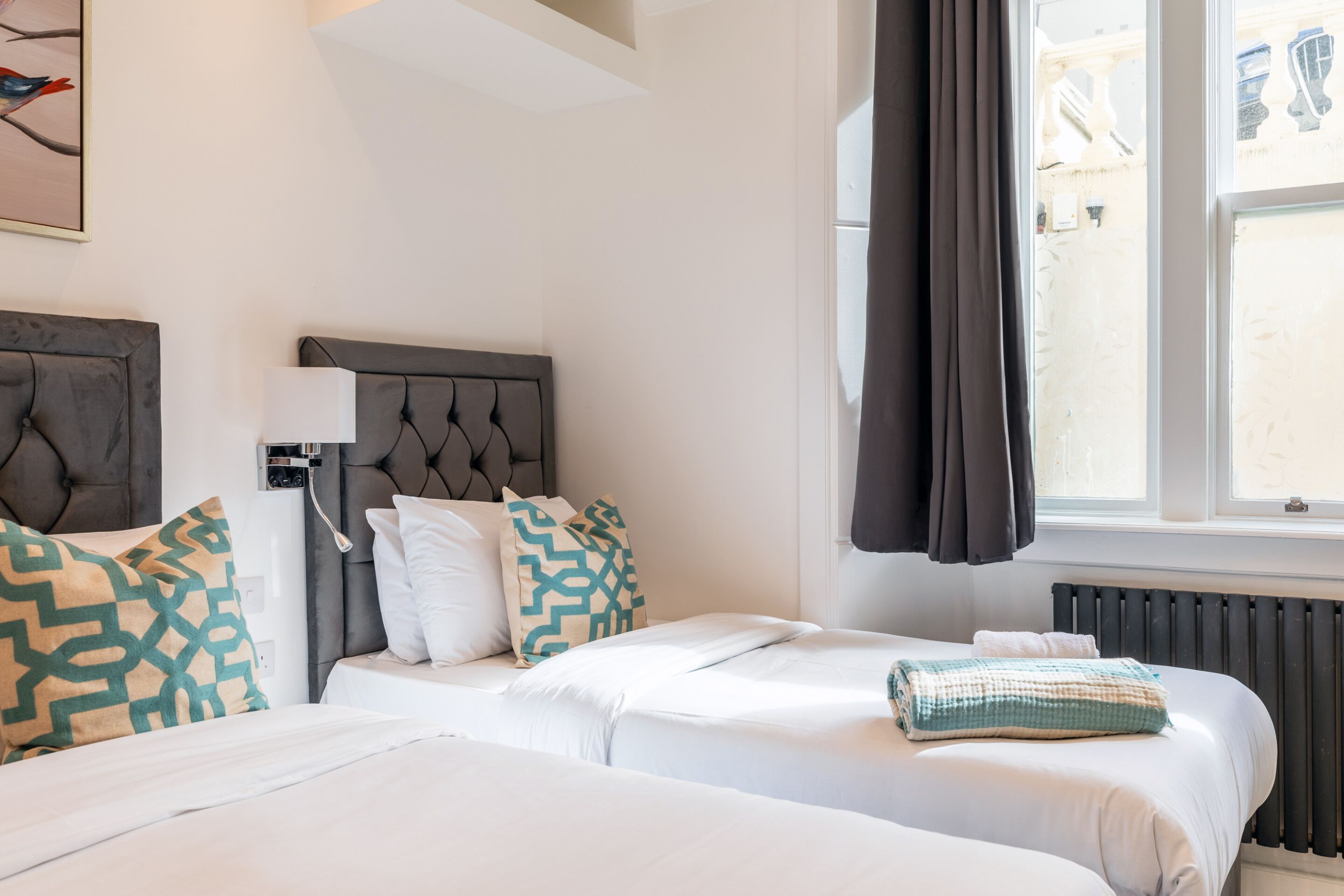 Habitación de hotel con dos camas, un radiador, una ventana con cortinas y un aplique de pared.