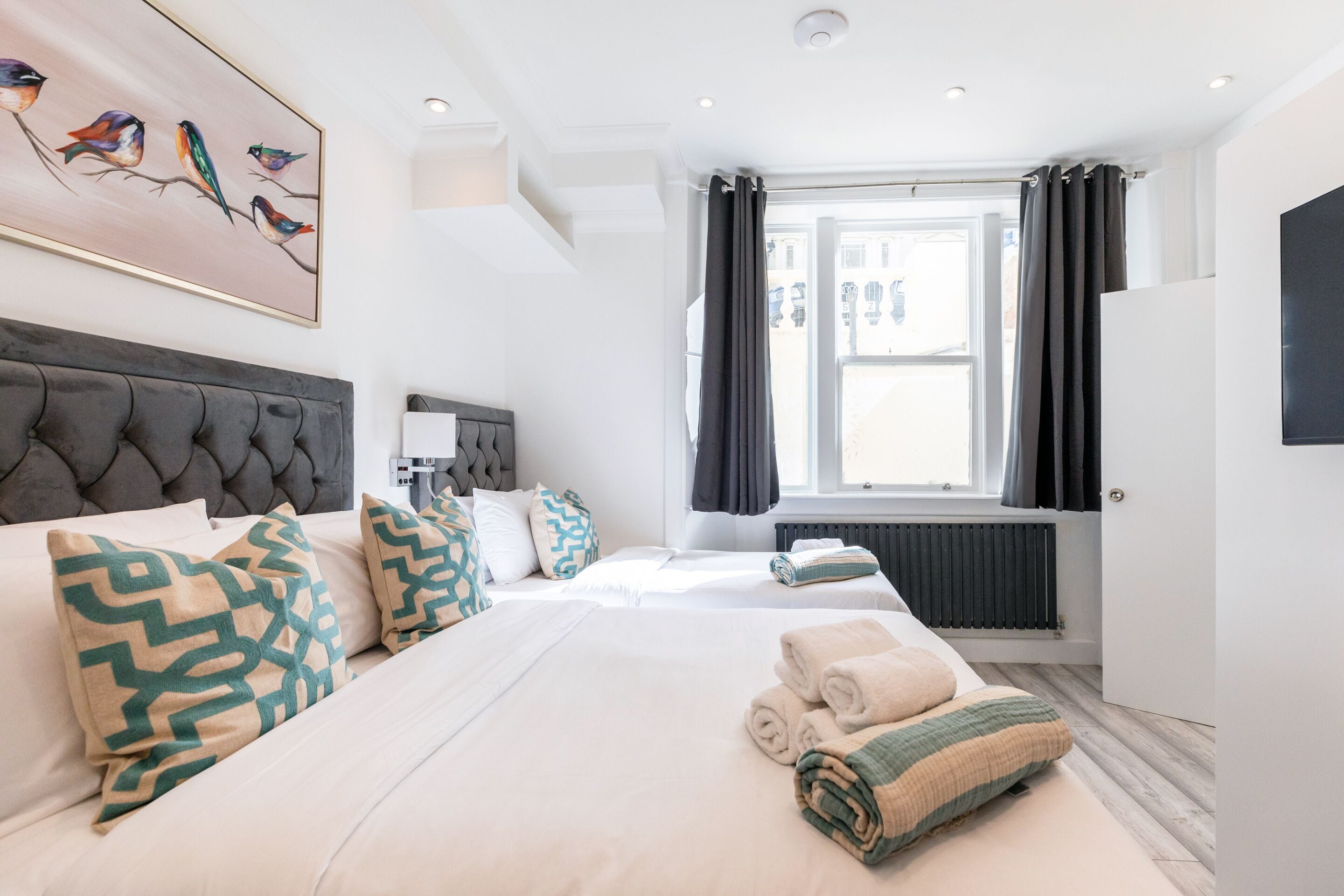 A hotel room with a large bed, two bedside tables, a headboard, a window with curtains, a radiator, and a painting of birds on the wall.