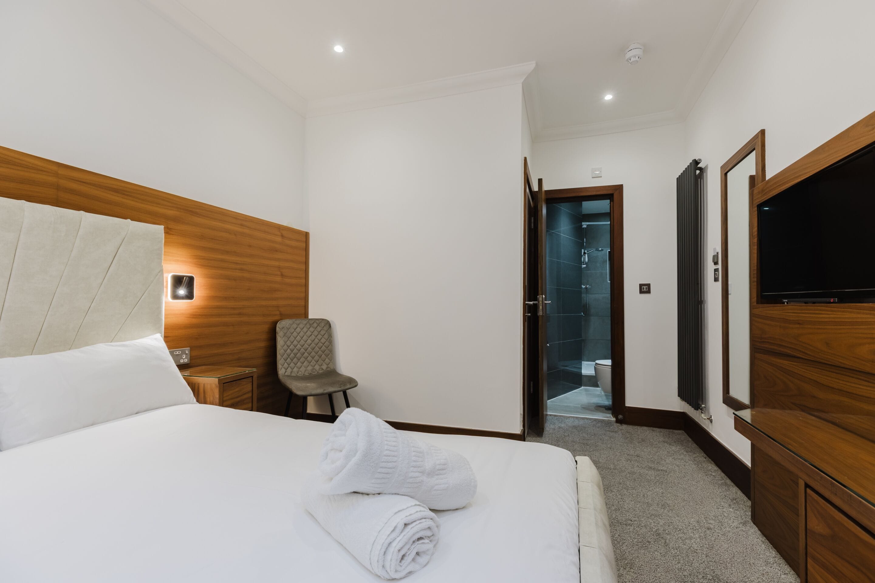 A hotel room with a bed, a chair, a TV, and a bathroom visible through an open door.