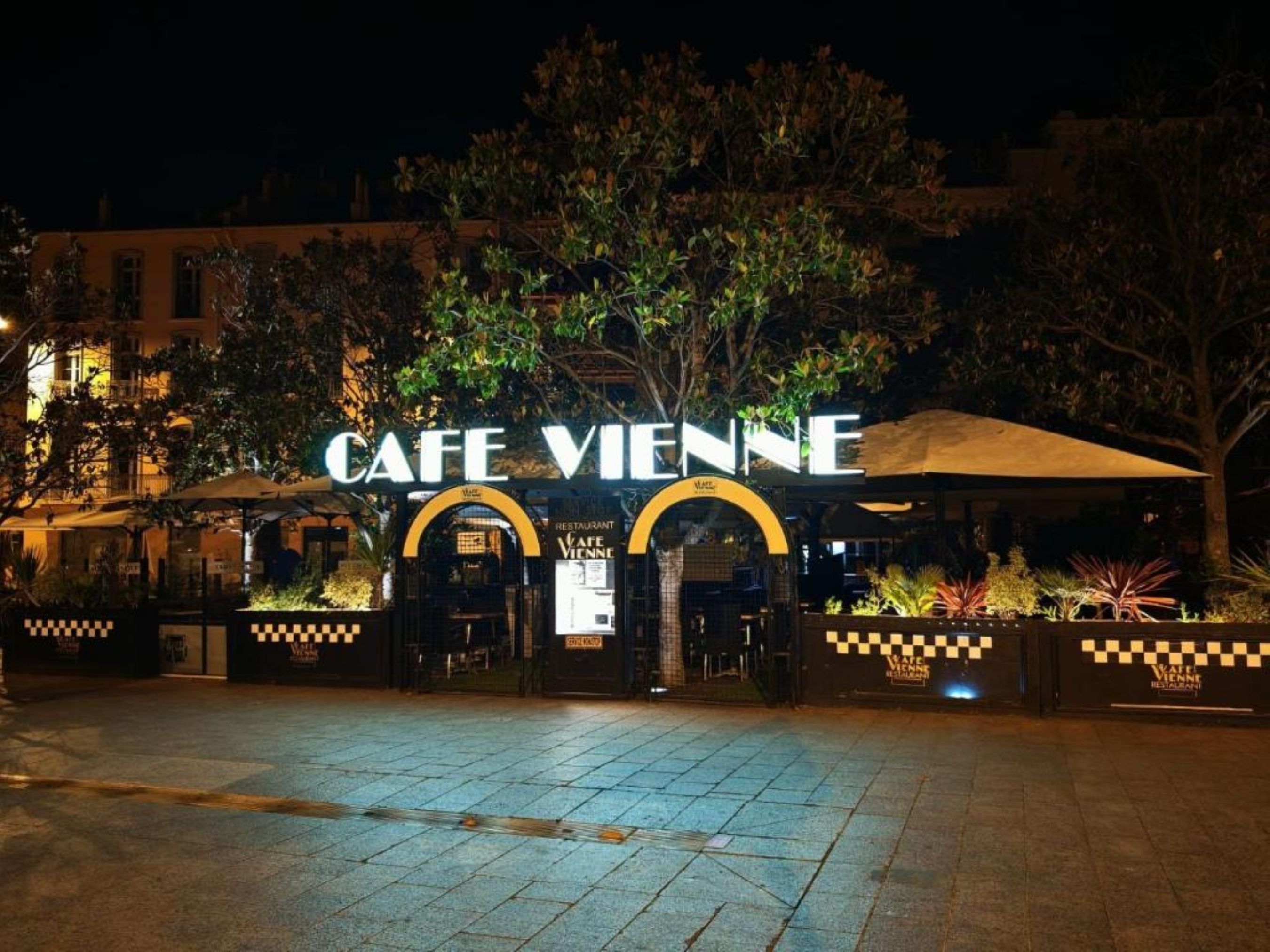 A nighttime view of a cafe with a prominent sign, outdoor seating, and a tiled pavement.
