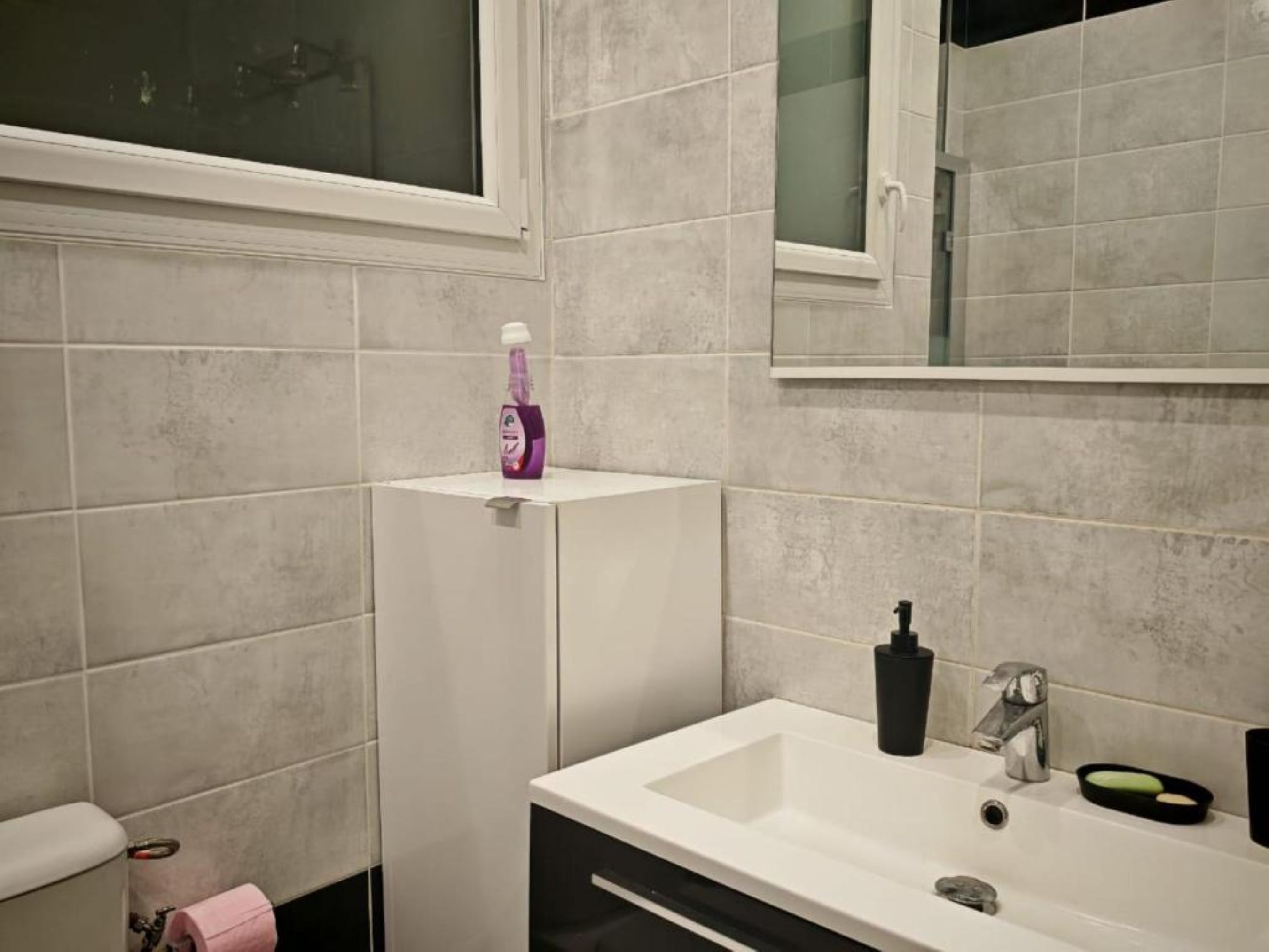 Un baño con un lavamanos blanco, un espejo encima y un estante con una botella.