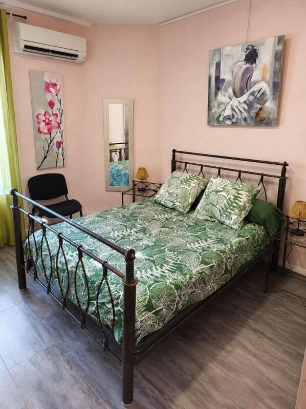 A bedroom with a bed, a chair, an air conditioner, a painting, and a mirror.