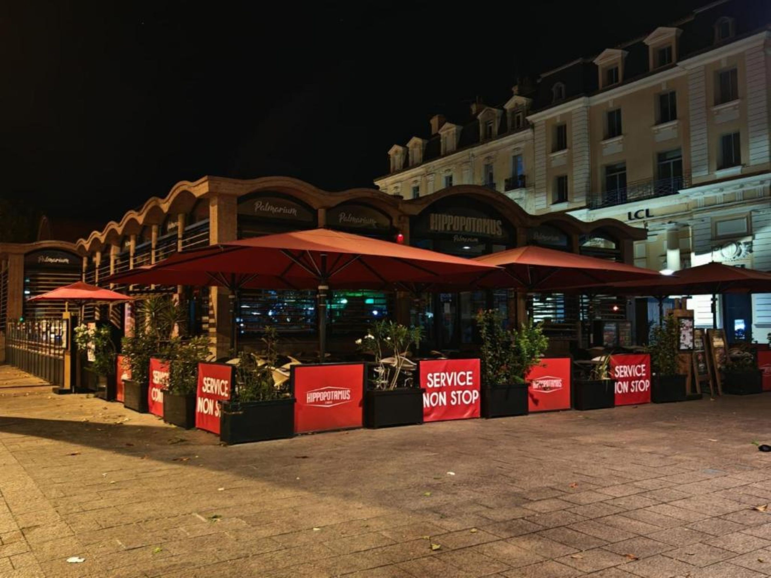 A restaurant with outdoor seating, red umbrellas, and signs indicating service availability.