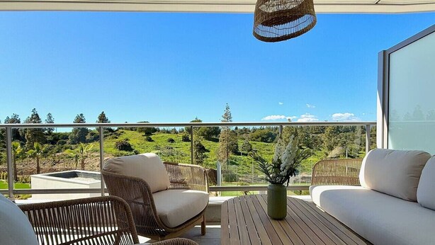Een balkon met rieten meubilair, een ronde houten tafel en uitzicht op een groen landschap.