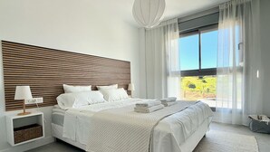 Een moderne slaapkamer met een groot bed, een nachtkastje met een lamp, uitzicht op groen en een hanglamp.