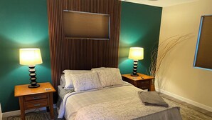 A neatly made bed with a patterned bedspread and pillows, flanked by two bedside tables with lamps, against a green wall with a wooden panel and a window with blinds.