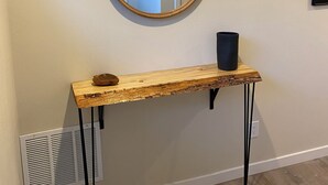 A wooden console table with a black vase and a round mirror above it, against a plain wall with a vent below.