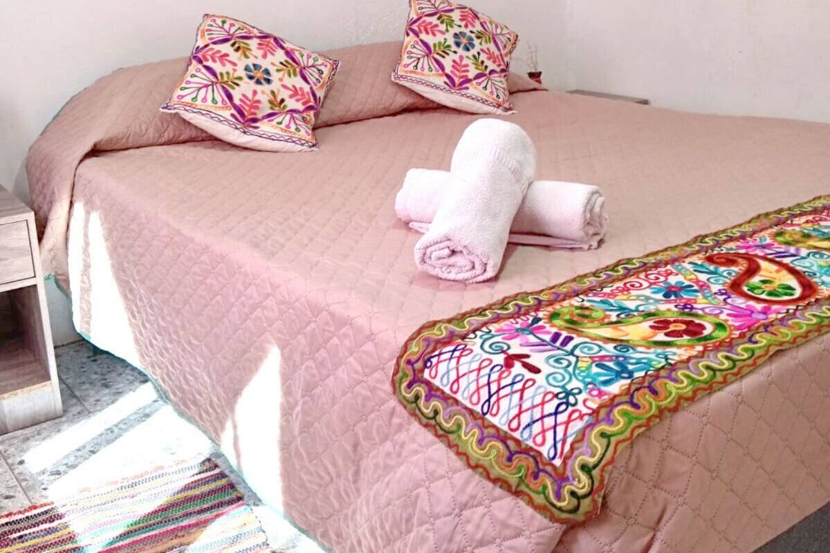 Ein Bett mit einer rosa Quilttdecke, zwei gemusterten Kissen, einem gerollten Handtuch und einer farbenfrohen Decke.
