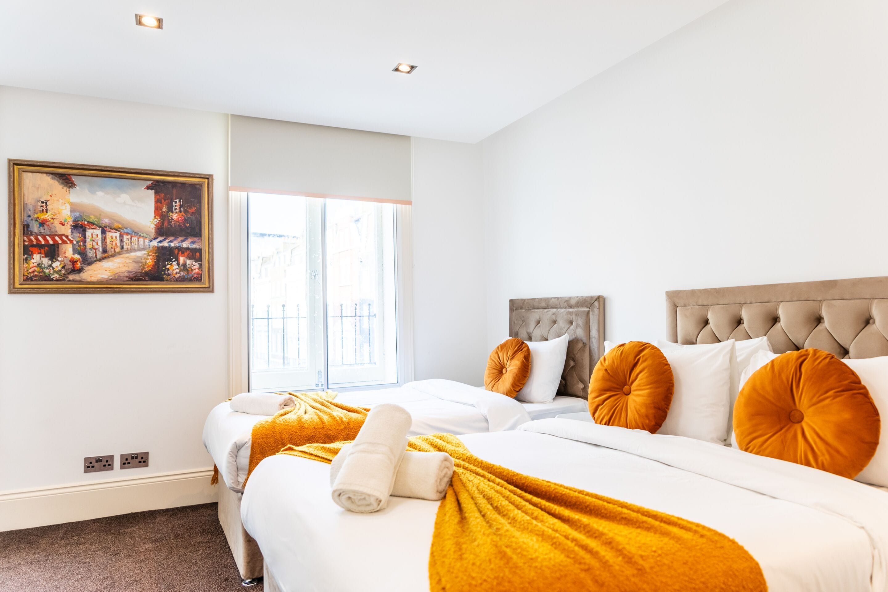 A hotel room with two beds, a painting on the wall, and a window with a view.