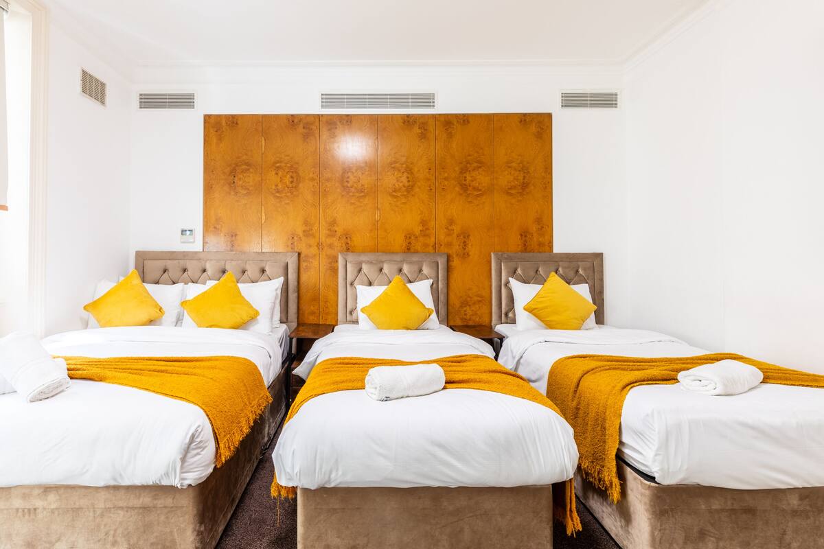 A hotel room with three beds, each with a tufted headboard and yellow pillows.