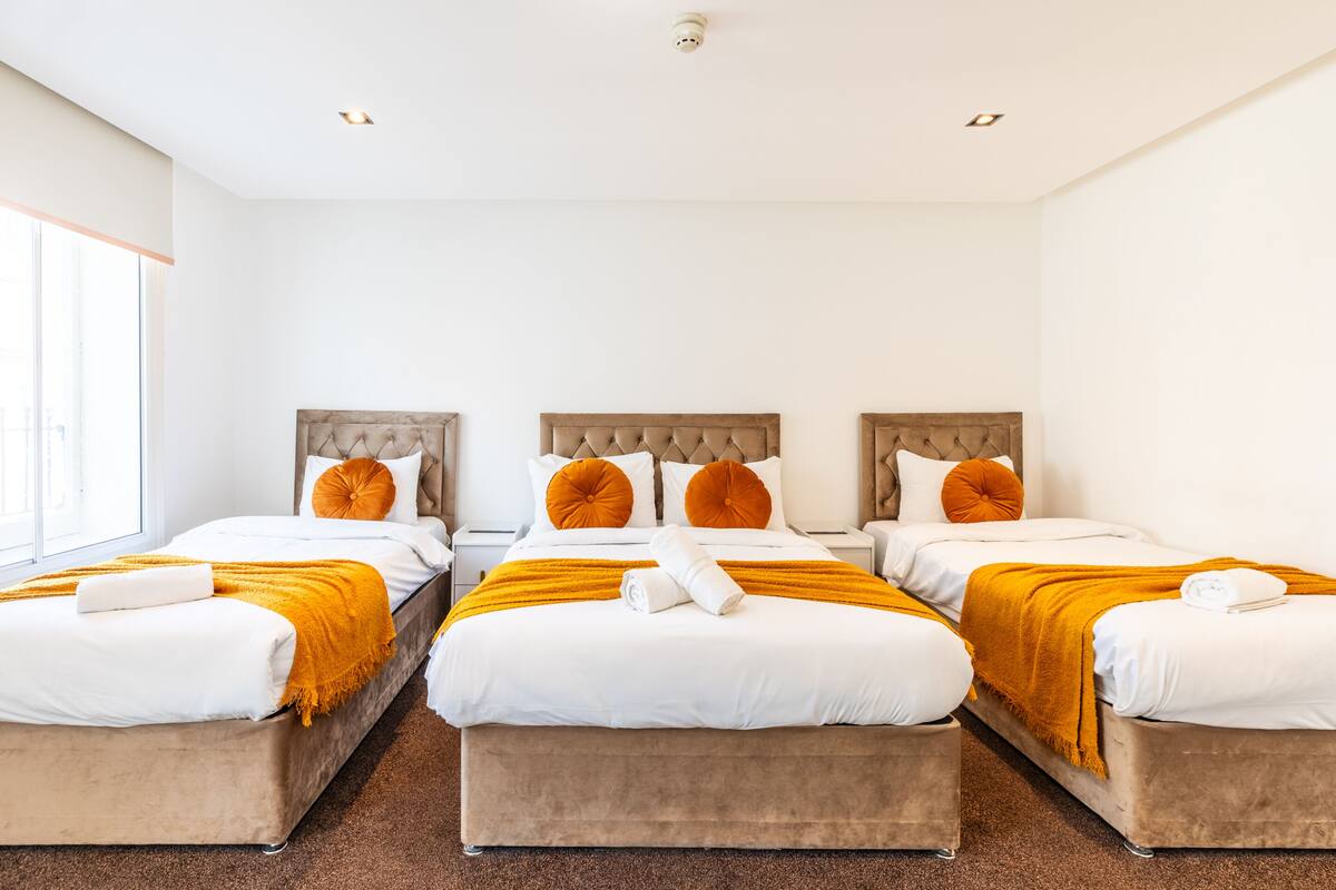 A hotel room with three beds, each with a tufted headboard, white bedding, and orange throw pillows.