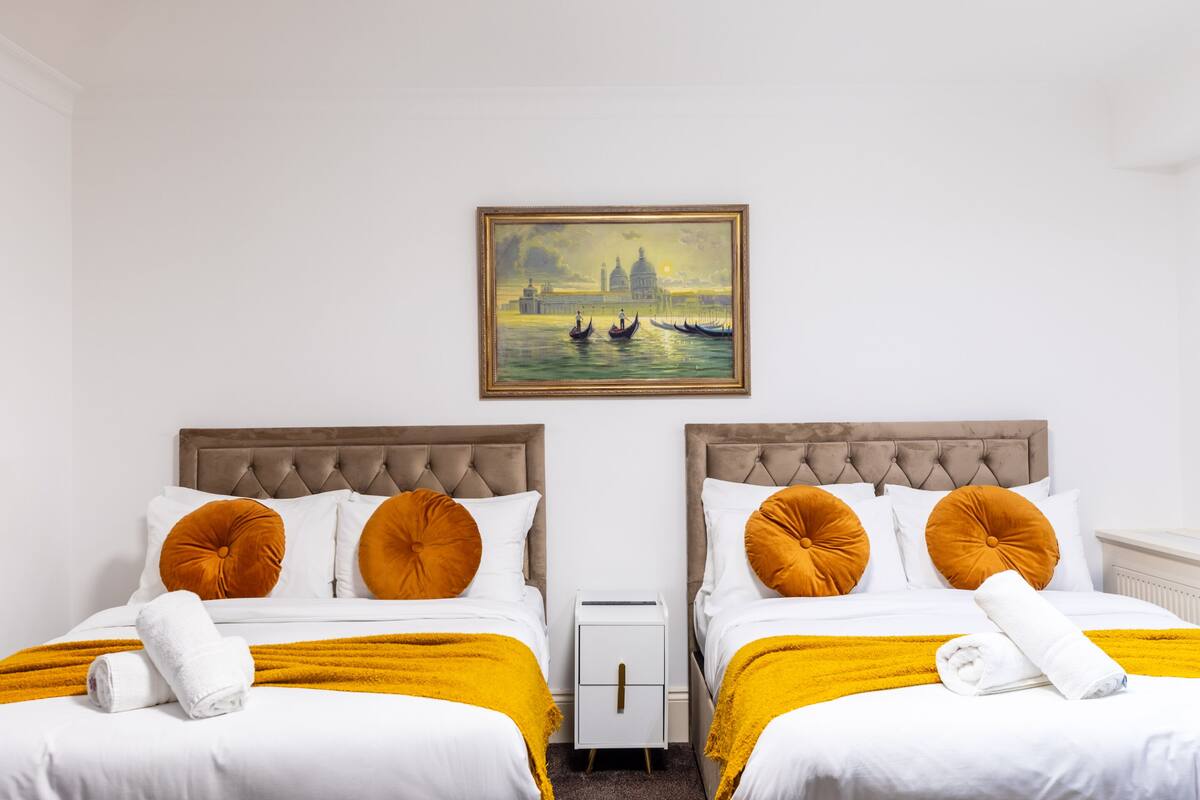 A hotel room with two beds, each with a tufted headboard, white bedding, and a yellow throw blanket. There is a painting of a cityscape on the wall above the beds.