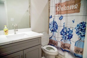 A bathroom with a white sink, a mirror, a toilet, and a shower curtain with a 'Welcome' sign. -  Charming 3-bedroom house with WiFi and AC in Houston - Unit #1 (Houston)
