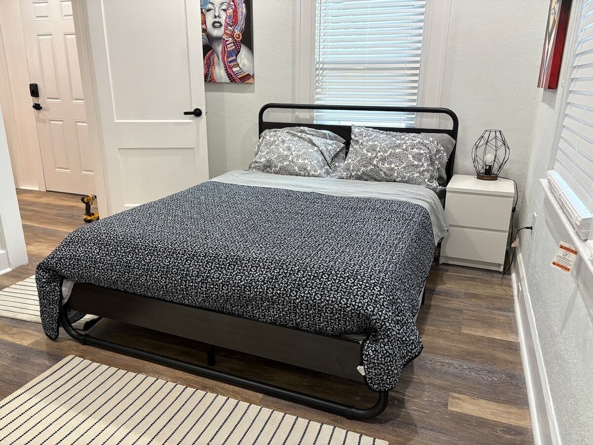 Ein Bett mit schwarzem Metallgestell und gemusterten Betttextilien, ein Nachttisch mit Lampe sowie eine Tür mit dekorativer Malerei.