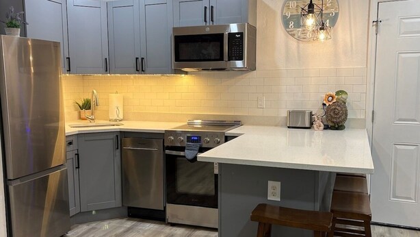 A modern kitchen with grey cabinets, stainless steel appliances, a white countertop, and a wooden stool. - St. Pete 5th Ave Oasis (St. Petersburg)