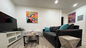 A modern living room with a flat-screen TV, a black sofa with teal cushions, a wooden coffee table, and two abstract paintings on the walls. - St. Pete 5th Ave Oasis (St. Petersburg)