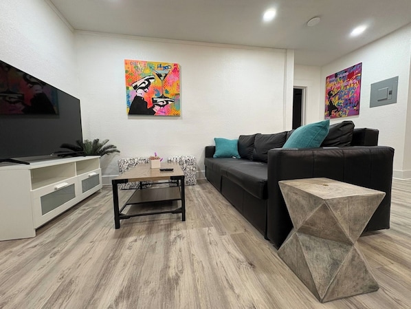 Ein modernes Wohnzimmer mit Flachbildfernseher, einer schwarzen Couch mit türkisfarbenen Kissen, einem Holztisch und zwei abstrakten Bildern an den Wänden.