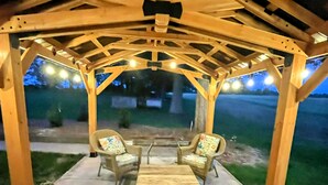 En täckt uteplats med en pergola av trä, fyra fåtöljer i rotting med blommiga kuddar och ett stort träbord i mitten.