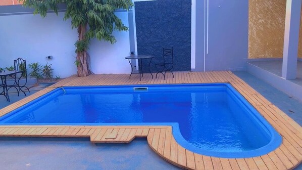 A rectangular swimming pool with a wooden deck, surrounded by a white wall and a dark textured wall. - Melia Beach (Mbour)