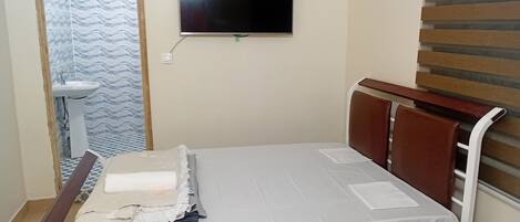 Ein Einzelbett mit weißer Matratze und Kissen, ein Holz-Kopfteil, ein an der Wand befestigtes Fernsehgerät und ein Badezimmer mit Waschbecken und gemusterten Fliesen.