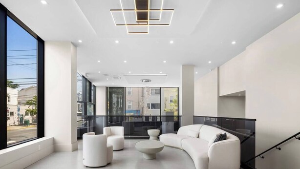A modern living room with a white sofa, two white armchairs, and a coffee table. There is a staircase with a black railing on the right.
