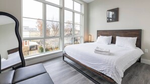 A bedroom with a large window, a bed with white bedding, a bedside table, and a bench.