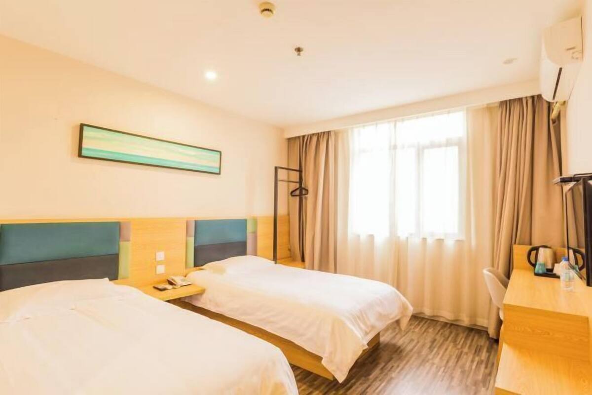 A hotel room with two beds, a desk, a chair, a window with curtains, and a painting on the wall.
