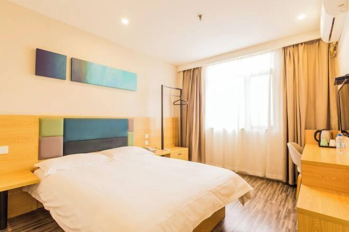 A hotel room with a bed, bedside table, a desk with a chair, a mirror, and a window with curtains.