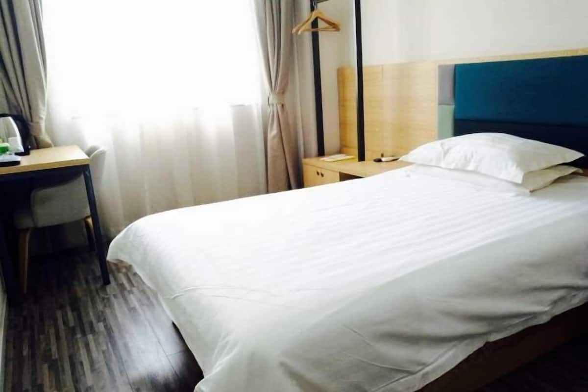 A hotel room with a bed, a desk, a chair, a nightstand, and a hanging coat rack.