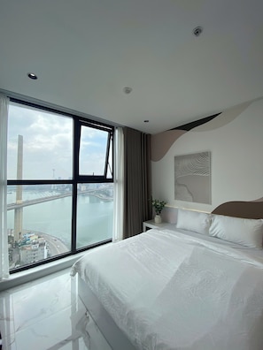 A hotel room with a large bed, a bedside table with a plant, a view of a bridge and water, and a modern ceiling design.
