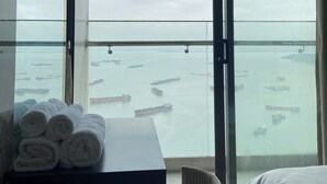 Ett hotellrum med ett stort fönster med utsikt över en hamn där flera fartyg ligger förtöjda. I rummet finns en säng, ett litet bord med hoprullade handdukar och en stol.