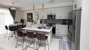 A modern kitchen with white cabinets, a central island with bar stools, a microwave, and a refrigerator. - Niagara Falls Area  Hidden Gem-Luxury, Cozy Canal-Side Home, Niagara Area's Hidden Gem- JNShortstay (Welland)