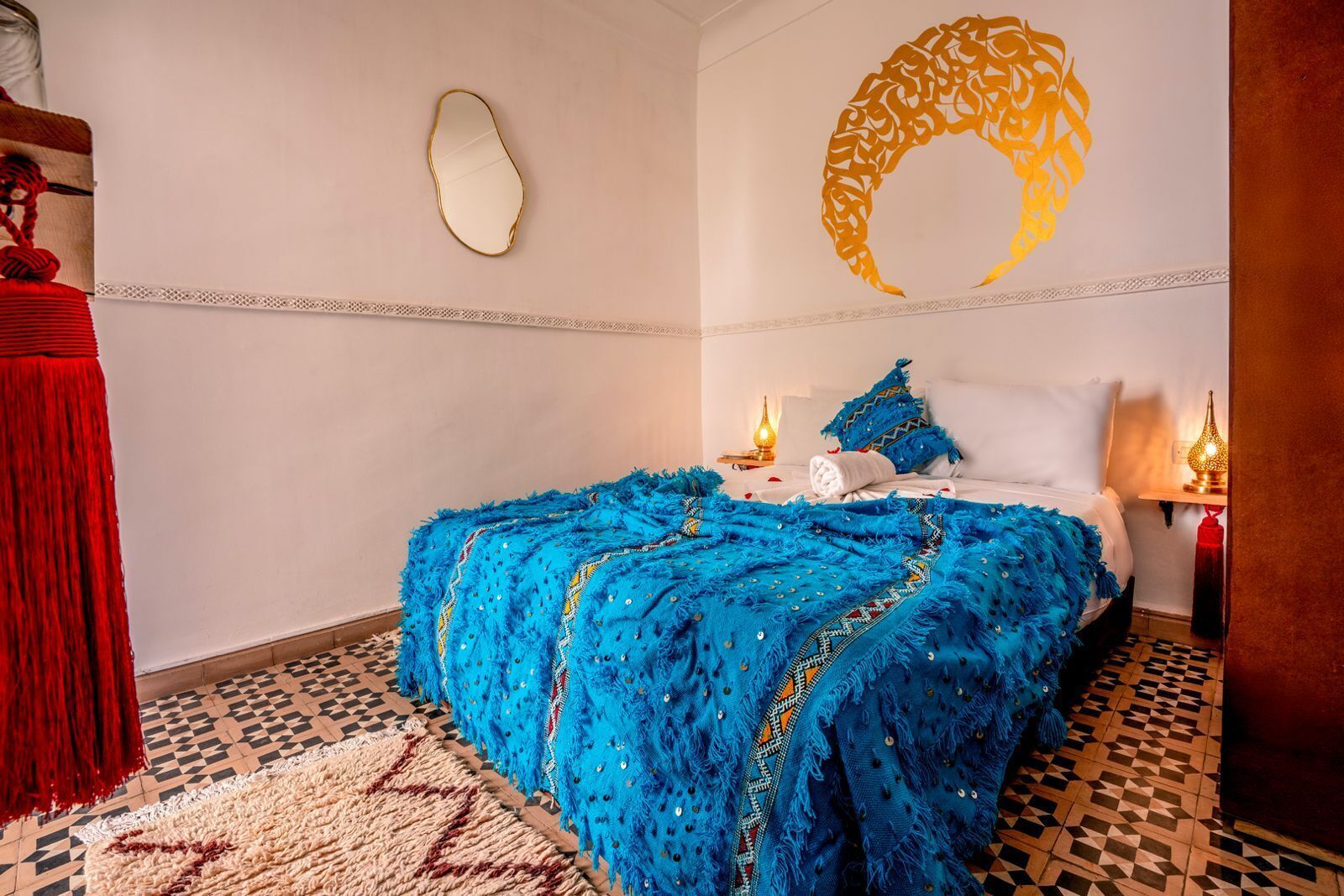 A bedroom with a bed covered in a blue, fringed blanket, a white pillow, and a decorative headboard. There is a small table with a lamp beside the bed, and a patterned rug on the floor.